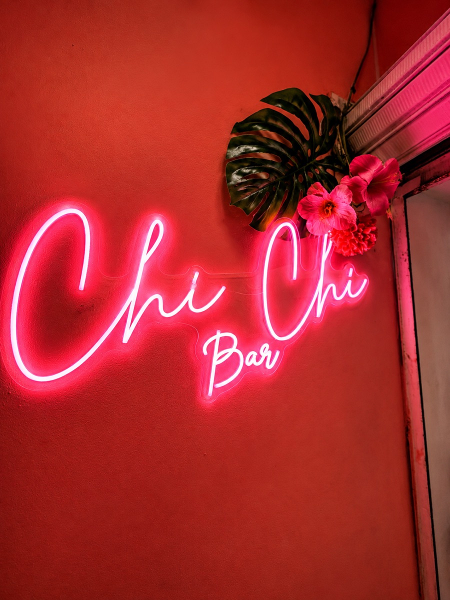 Letrero de neón rosado 'Chi Chi Bar' sobre pared coral, con monstera y flores tropicales